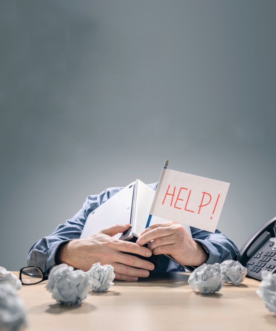 Mann der sich mit Oberkörper auf Schreibtisch legt und den Kopf hinter aufgeklapptem Buch verbirgt und in der Hand eine Fahne mit Aufschrift HELP hält