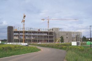 Im Bau befindliches Firmengebäude auf das eine Straße zugeht umrandet von grün mit Baukränen im Vordergrund
