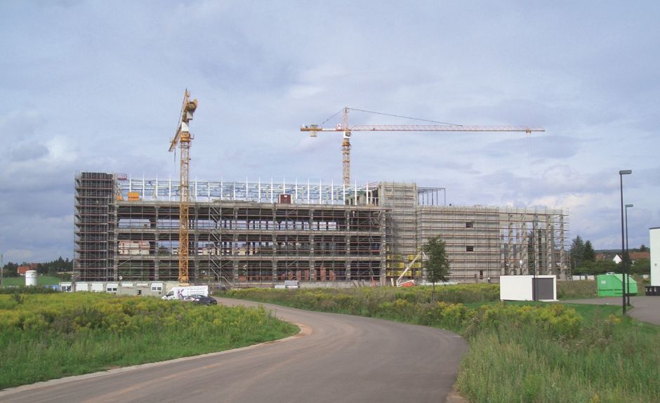 Im Bau befindliches Firmengebäude auf das eine Straße zugeht umrandet von grün mit Baukränen im Vordergrund