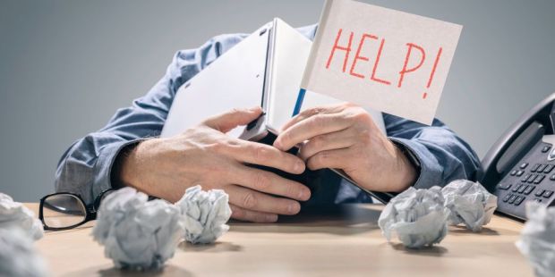 Mann der sich mit Oberkörper auf Schreibtisch legt und den Kopf hinter aufgeklapptem Buch verbirgt und in der Hand eine Fahne mit Aufschrift HELP hält