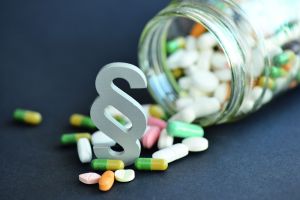 Pillen die aus Glas fallen mit Paragraphensymbol; Erfahren Sie, was GMP bedeutet, welche Ziele verfolgt werden und wie die Standards in Pharma-, Lebensmittel- und Kosmetikindustrie umgesetzt werden.
